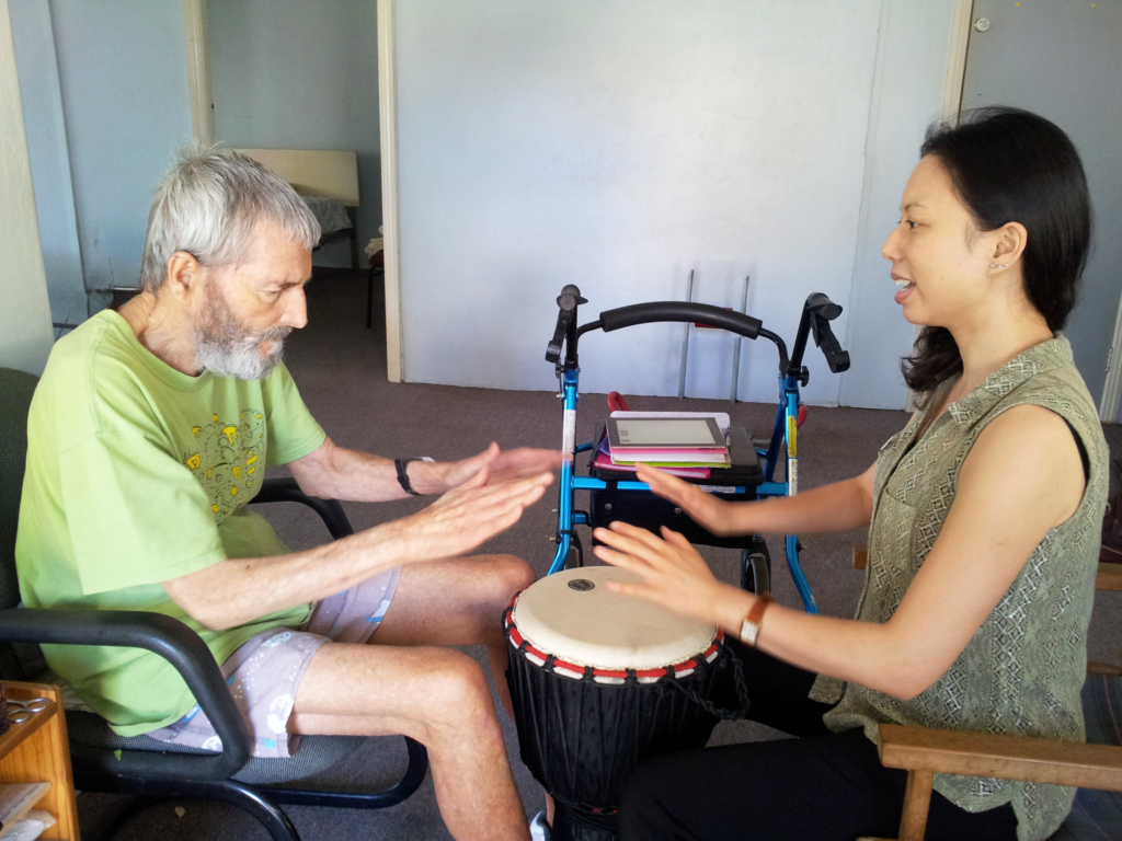 Two adults seated opposite each other clap on a djembe drum between them in a therapy room, with a walker in the background.