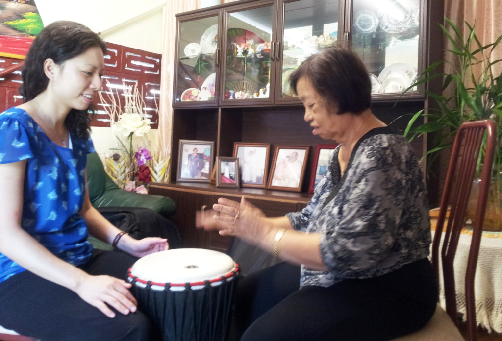Two women sit facing each other while one plays a djembe drum and the other claps, smiling in a living room setting with cabinets and photos behind them.